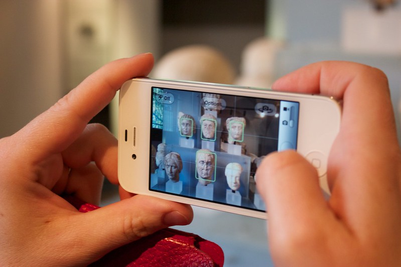 a pair of caucasian hands holding a white iphone in horizontal orientation. It is in photo mode and is taking a picture of roman bust sculptures. The green face-tagging boxes are around the faces of the sculptures
