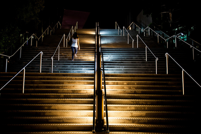 a picture looking up a big flight of metal stairs at night, empty but for one woman walking up them