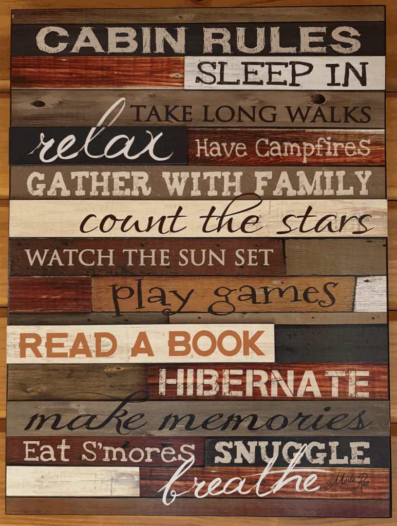 This is a pic of a motivational sign that is seemingly made of wooden planks. In all different fonts and colors it reads: "Cabin Rules: Sleep in, Take long walks, relax, have campfires, gather with family, count the stars, watch the sun set, play games, read a book, hibernate, make memories, eat s'mores snuggle, breathe"
